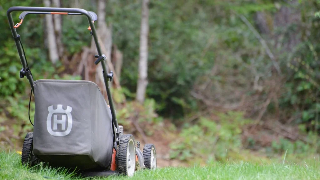 Husqvarna push mower with grass catcher parked on a green lawn at the edge of a forest